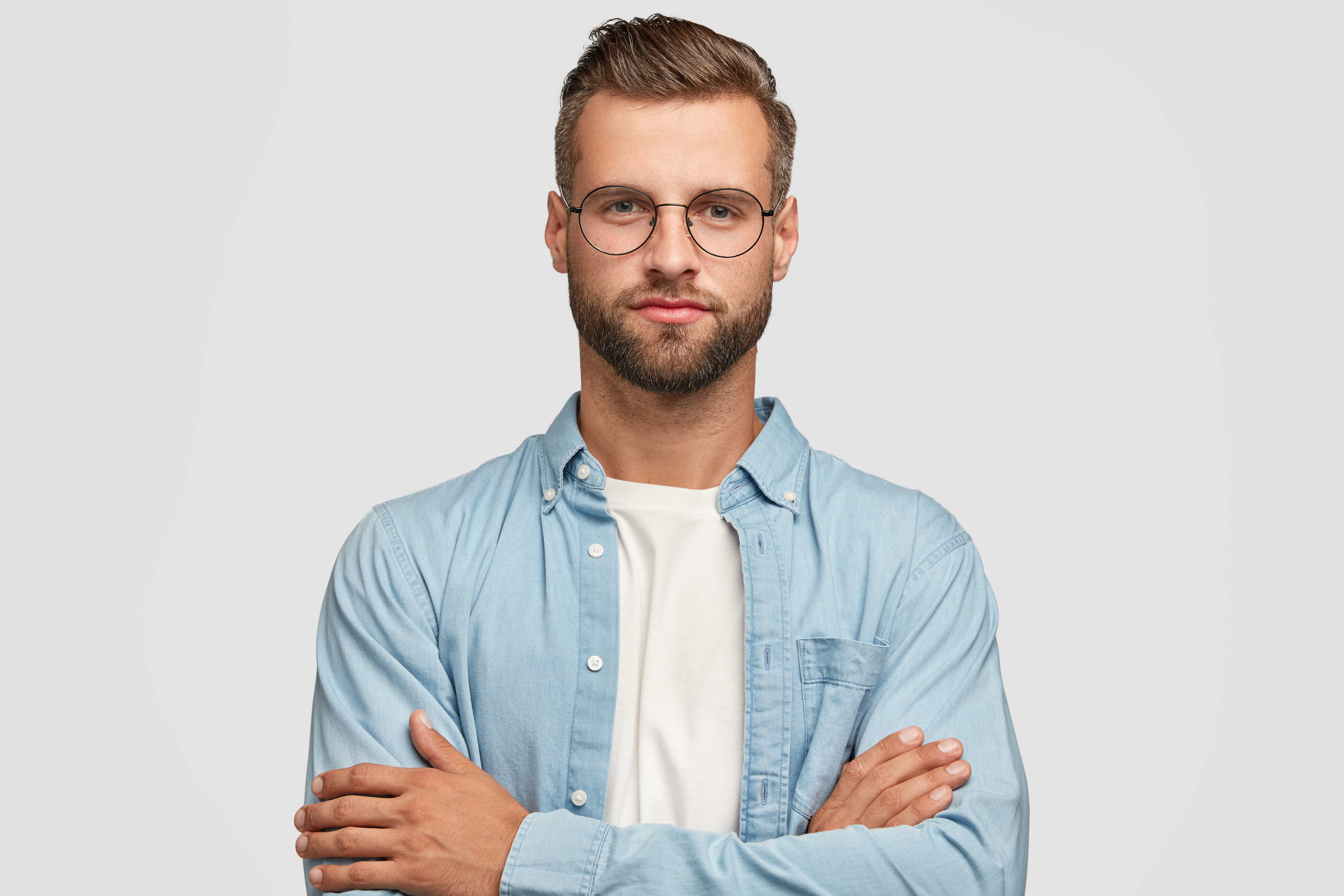 Horizontal shot of handsome self confident businessman or designer, stands crossed hands against white background, wears casual shirt, poses against white background